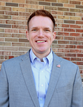 Man in a light blue suit stands in front of a brick wall.